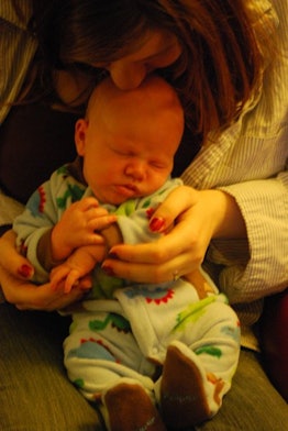A woman in postpartum holding her newborn dressed in a onesie on her lap and kissing the baby's fore...