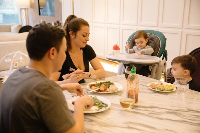 A mother, father, son, and a baby sitting in a baby gear feeding chair having a meal in a white kitc...