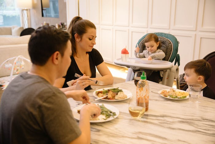 A mother, father, son, and a baby sitting in a baby gear feeding chair having a meal in a white kitc...