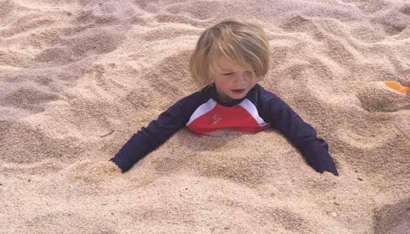 A little blonde-headed boy buried in beach sand, playing