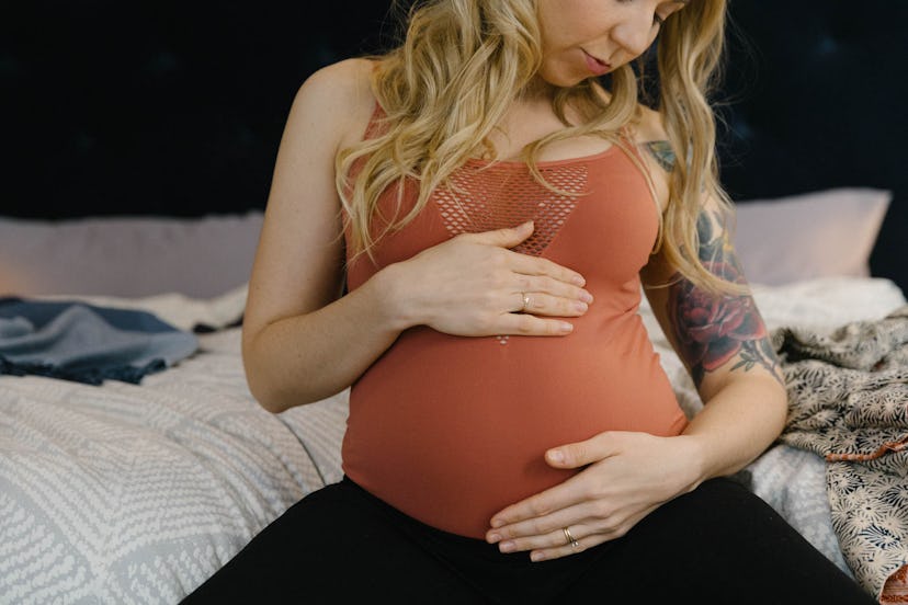 A blonde pregnant woman holding her belly while sitting on a bed