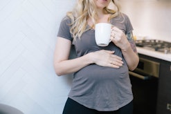 A pregnant woman in a grey t-shirt feeling her belly while holding a cup