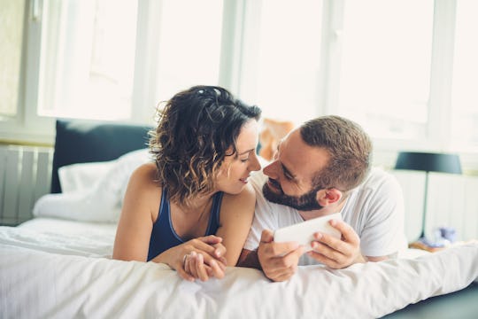 A man holding his phone while leaning toward his partner lying next to him on a bed