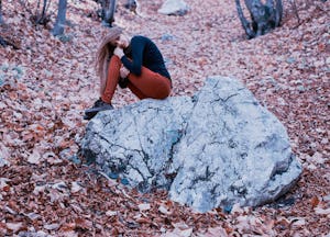 A woman with bipolar disorder sitting on a bolder in a forest, leaning her head against her knees 