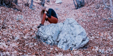 A woman with bipolar disorder sitting on a bolder in a forest, leaning her head against her knees 