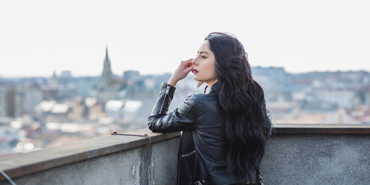 A dark-haired woman leaning against a fence of a building rooftop, being alone to help her depressio...