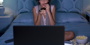 A young woman sitting on her bed, eating popcorn while watching TV
