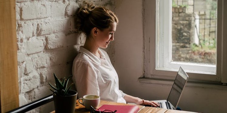 A woman who wants to reduce stress at work smiling and sitting next to a window while using her lapt...