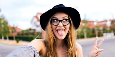 A woman in a black floppy hat, glasses, and a sweater sticks out her tongue and makes a peace sign w...