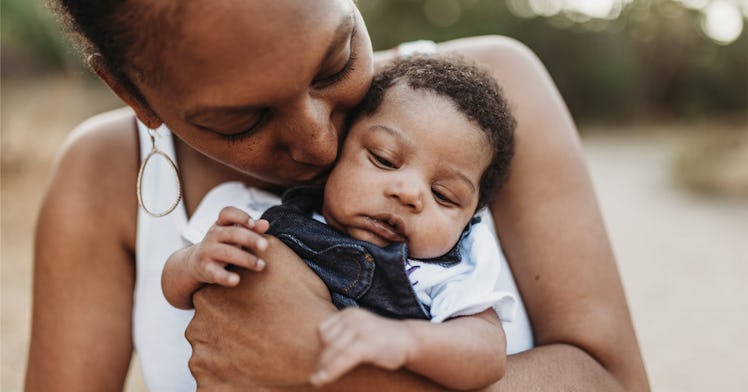 mother wearing white tank top holding and kissing newborn in blue overalls on mothers rday