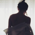 A woman standing in front of a window with curtains, back to camera taking off her shirt