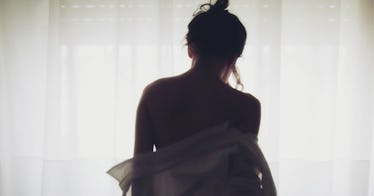 A woman standing in front of a window with curtains, back to camera taking off her shirt