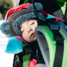A baby in winter clothes napping in a backpack on a parent's back outside, with snow in the backgrou...