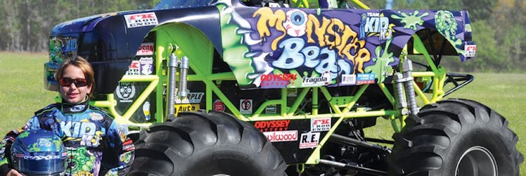 A little boy in a leather jacket, glasses and a helmet next to a multi-colored Mini Monster Truck