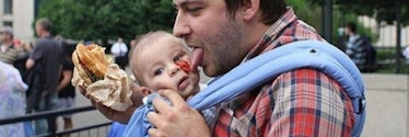 A dad holding his son and eating a hamburger with ketchup spilled over the toddler's face