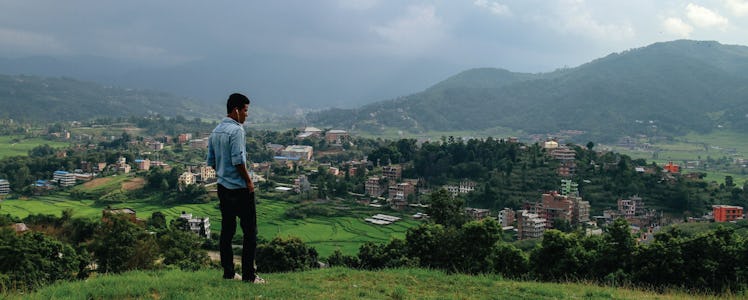 A man standing on a hill overlooking a small town surrounded by greenery and mountains