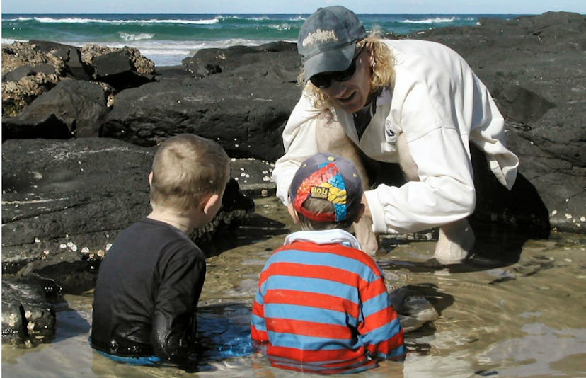 A man at the seashore sitting in front of two toddlers talking to them 