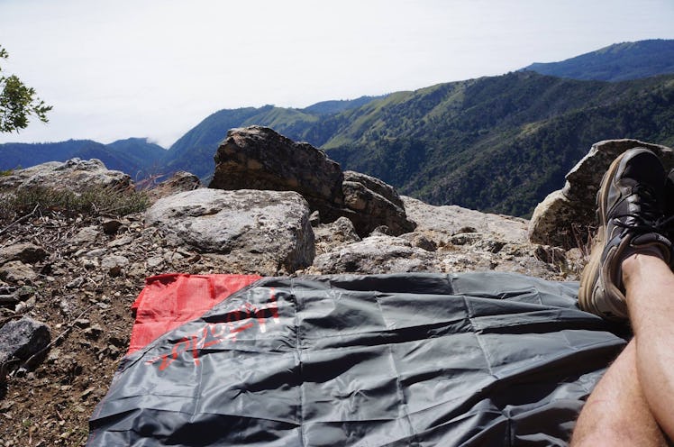 A man sitting on a blanket that can fit in his pocket on top of a mountain site during the day