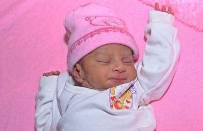 A baby in a white shirt and pink hat laying down on a pink blanket 