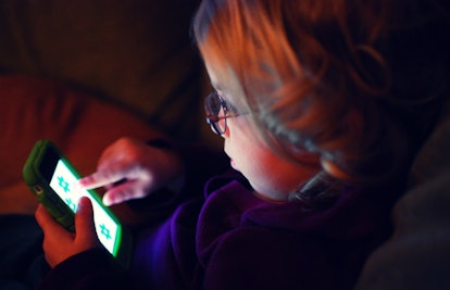 A little digitally savvy girl wearing glasses sitting in a dark room and using her smartphone who ha...