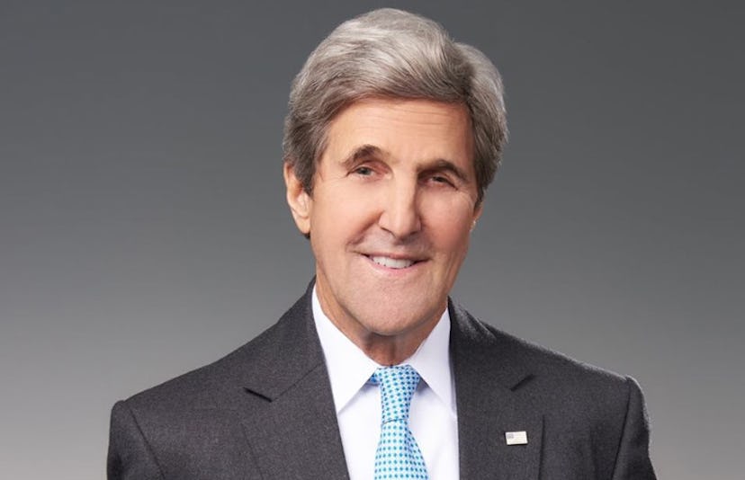 A closeup of John Kerry in a grey suit, white button-up and light blue tie with a flag of the USA pi...