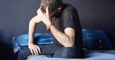 Sexually frustrated man sitting on the edge of a bed cradling his head wearing jeans and a t-shirt