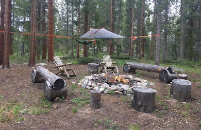 A campsite with logs and chairs around a campfire and a tree tent in the background