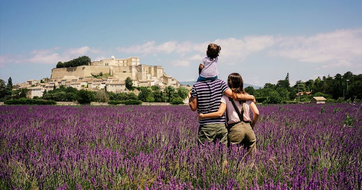 A mom and dad with a baby on his shoulders stand in a field of purple flowers, looking at architectu...