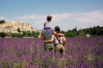 A mom and dad with a baby on his shoulders stand in a field of purple flowers, looking at architectu...