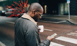A man stands on the sidewalk and stares down at his smartphone, one of the main causes of tech neck.