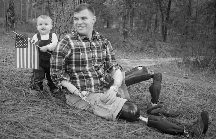 Travis Mills, purple heart winner sits on the ground next to his son who is holding a small american...