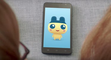 A Tamagotchi app opened on a smartphone, and two people looking at it from the distance.