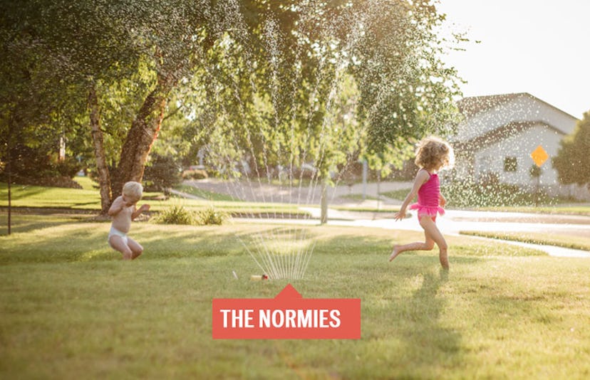 Two children running beneath the spray of sprinklers in their yard with a sign that reads The Normie...