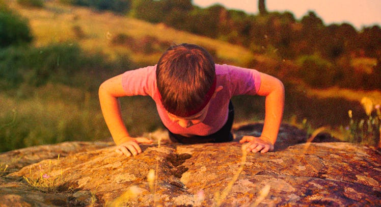 A kid in a pink shirt doing push ups on a rock next to a hill