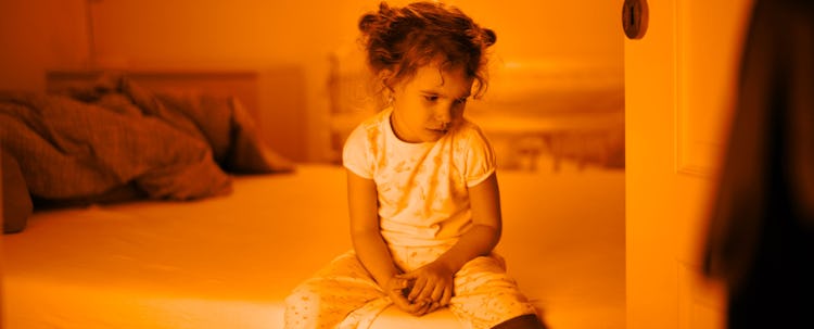 An angry little girl sitting on the edge of her bad after being disciplined by her parents using a b...