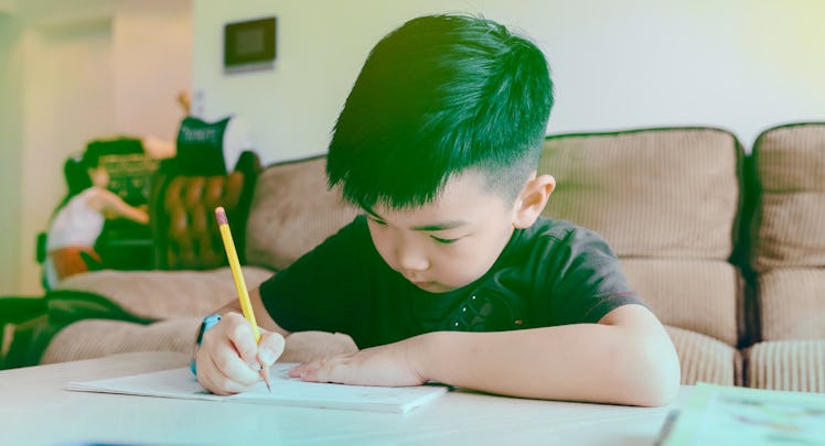 A boy writing his homework, sitting behind a table, while a woman is cleaning up in the background. 