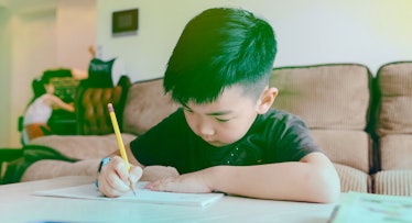 A boy writing his homework, sitting behind a table, while a woman is cleaning up in the background. 