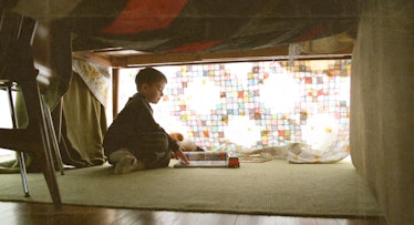 A child sitting under blankets in an imaginary fort reading a book which is in front of him