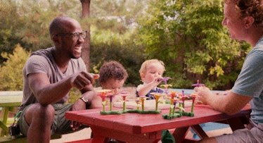 Two shy dad friends sitting outside at a picnic with their children laughing and playing with toys