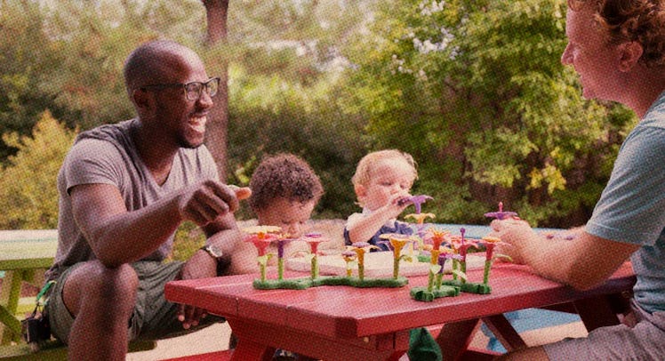 Two shy dad friends sitting outside at a picnic with their children laughing and playing with toys