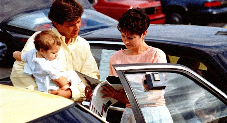 A man holding a baby and his wife standing next to him showing him newspapers on a parking lot befor...