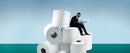 A man reading a newspaper, dressed in a suit, sitting atop a pile of giant rolls of toilet paper.