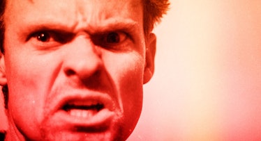 A very angry man staring straight ahead, he is bathed in a red light.