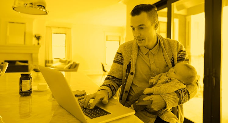 A new dad holding his baby and typing on a laptop and using social media in his kitchen