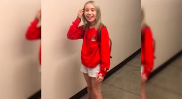 Lil Tay standing in a red sweater and white shorts while moving her hair away from her face with her...