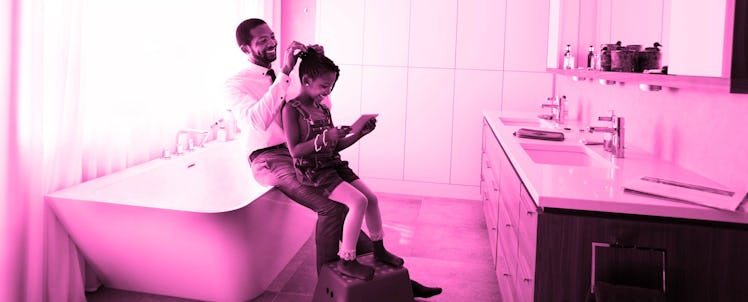 A smiling dad sitting on the edge of a bathtub braiding his daughter's hair