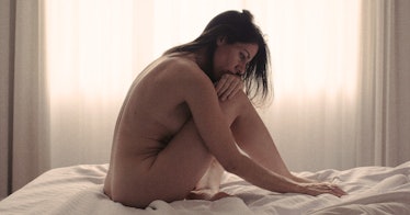 A naked, curled-up brunette woman sitting on her bed struggling with her postpartum body