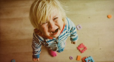 A crying blonde toddler in a white-blue striped onesie looking up with toys around his feet