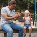 A dad and child on a bench at a playground clapping hands.