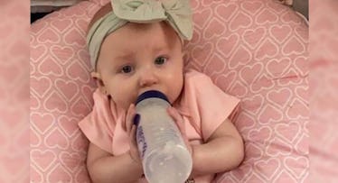 A baby in a pink shirt and a beige bow on her head drinking milk from a bottle